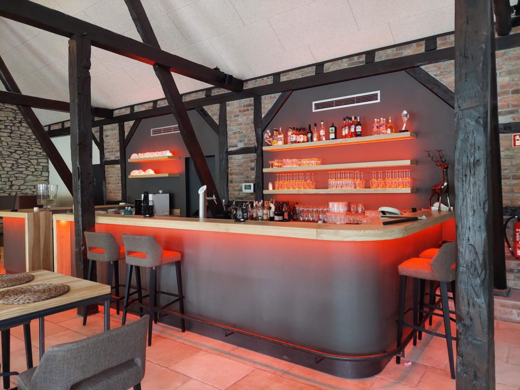 Alt-Text: Moderne Bar mit Holz- und Steinarchitektur, beleuchtet in warmen Rottönen. Im Hintergrund sind Regale mit verschiedenen Spirituosen und Gläsern zu sehen. In der Mitte der Bar stehen hohe Stühle, und die Theke hat eine helle Holzoberfläche. Die Umgebung wirkt gemütlich und einladend.