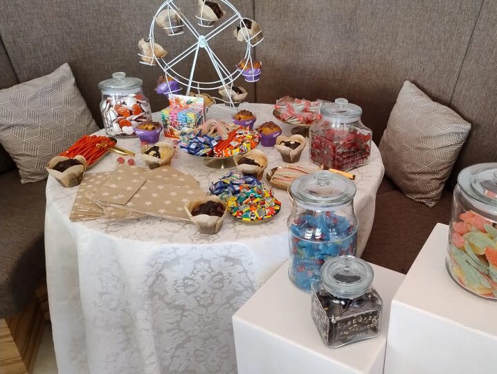 Ein festlich gestalteter Naschtisch in einer gemütlichen Sitzecke. Auf dem mit weißer Spitzentischdecke gedeckten runden Tisch stehen viele bunte Süßigkeiten in Gläsern, Schälchen und Muffinförmchen. In der Mitte befindet sich ein kleines weißes Dekorationsriesenrad, das ebenfalls mit Süßigkeiten gefüllt ist. Zwei zusätzliche Bonbongläser stehen auf weißen Podesten rechts neben dem Tisch.