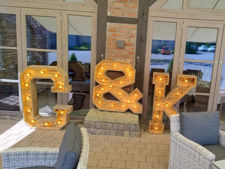 Große, beleuchtete Holzbuchstaben ‚G & K‘ stehen dekorativ vor einer Fensterfront mit Fachwerk und Backstein. Die Buchstaben sind mit kleinen Glühbirnen versehen und leuchten warm. Sie stehen auf einer gepflasterten Terrasse zwischen Sitzmöbeln aus Rattan – vermutlich als Hochzeitsdekoration