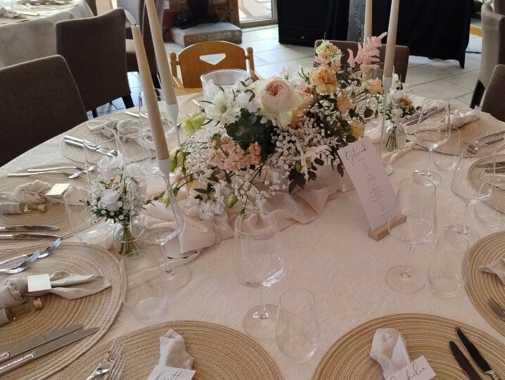 Festlich gedeckter runder Tisch mit hellen Platzsets, Stoffservietten, Besteck, Gläsern und Namenskarten. In der Mitte steht ein längliches Blumenarrangement mit weißen und zartrosafarbenen Blüten, Schleierkraut und Eukalyptus, dekoriert mit weichem Stoff. Zwei helle Stabkerzen und eine Menükarte ergänzen die elegante Tischdekoration. Im Hintergrund steht ein Kinderhochstuhl.