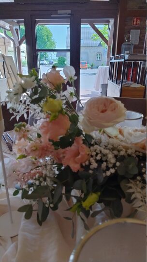 Ein Blumengesteck mit zarten rosa Rosen, weißem Schleierkraut, kleinen gelben Blüten und Eukalyptus steht auf einem festlich gedeckten Tisch. Im Hintergrund ist ein geöffneter Glaseingang zu sehen, durch den man nach draußen auf einen sonnigen Hof mit Kopfsteinpflaster und Gebäuden blickt.