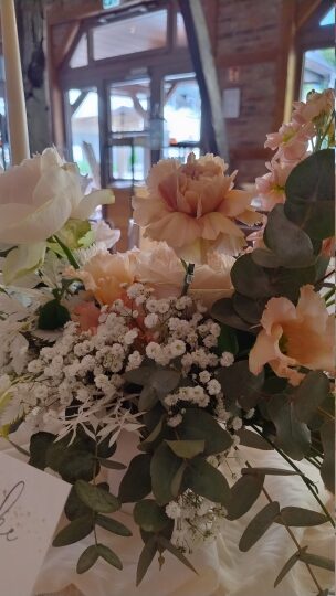 Nahaufnahme eines eleganten Blumenarrangements auf einem festlich gedeckten Tisch. Zu sehen sind zartrosafarbene Pfingstrosen, weiße Rosen, Schleierkraut und Eukalyptusblätter. Im Hintergrund erkennt man unscharf den Eingang eines rustikalen Veranstaltungsraums mit großen Glasfenstern und Holzbalken
