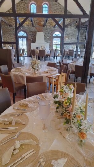 Eleganter, festlich gedeckter Veranstaltungsraum mit runden Tischen, weißen Tischdecken, beigefarbenen Platzsets und cremefarbenen Stoffservietten. Die Tische sind mit Blumenarrangements in Pastelltönen und langen, schmalen Kerzen dekoriert. Im Hintergrund sind hohe Fenster, eine Steinwand und eine Kaminverkleidung in einem rustikal-modernen Ambiente mit offenem Gebälk zu sehen.