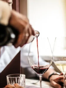 "Ein eleganter Moment in einem Restaurant, bei dem ein Kellner Rotwein in ein großes Weinglas einschenkt. Im Hintergrund ist ein weiteres Glas Weißwein zu sehen, was die Szene zu einem stilvollen, gastronomischen Erlebnis macht. Der Fokus liegt auf dem Fließen des Weins, das in der warmen, einladenden Atmosphäre des Lokals eine gehobene Stimmung erzeugt