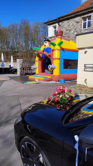 Im Vordergrund ist die Motorhaube eines schwarzen Autos mit Blumenschmuck und weißem Schleifenband zu sehen – vermutlich ein Hochzeitsauto. Im Hintergrund steht eine bunte Hüpfburg mit Piratenmotiv vor einem Gebäude mit Natursteinfassade. Links daneben befinden sich weiße Sonnenschirme und Tische eines Außenbereichs. Es ist sonniges Wetter, die Bäume im Hintergrund sind noch kahl.