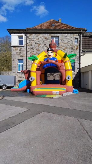 Bunte Hüpfburg mit Piratenmotiv, aufgebaut auf einem gepflasterten Innenhof vor einem Gebäude mit Natursteinfassade und rotem Ziegeldach. Die Hüpfburg ist in leuchtenden Farben gestaltet, mit aufblasbaren Palmen, einem Piratenkopf mit Hut und zwei großen Augen an den Seiten. Der Himmel ist blau mit vereinzelten Wolken.
