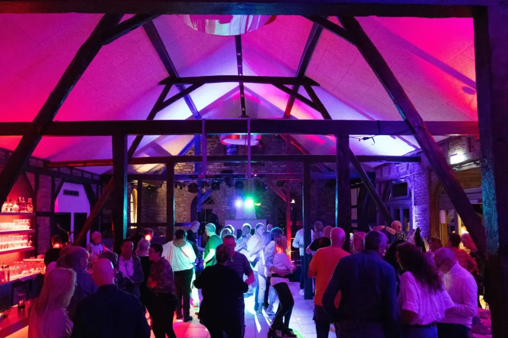 Eine lebhafte Tanzveranstaltung in einem stilvollen Veranstaltungsraum mit einem hohen, holzverkleideten Gewölbe. Die multicolor Beleuchtung schafft eine festliche Atmosphäre, während Gäste in eleganter Kleidung auf der Tanzfläche aktiv sind. Im Hintergrund sind eine Bar und weitere Gäste sichtbar, die sich unterhalten.