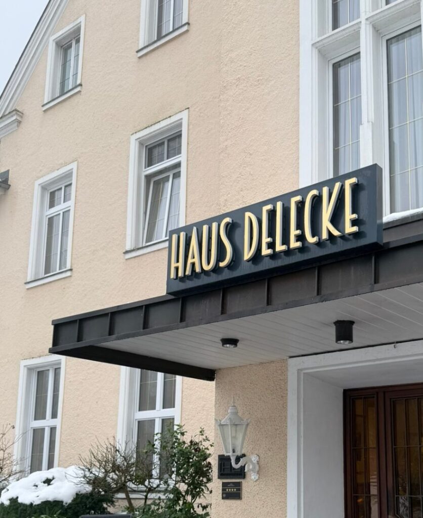Nahaufnahme des Eingangsbereichs von 'Haus Delecke', zu sehen an einem eleganten Schild über der Tür. Das Schild trägt den Namen in stilvollen, goldenen Buchstaben auf dunkelblauem Hintergrund, und wird von klassischen weißen Fenstern und einer dekorativen Laterne begleitet. Im Vordergrund ist leicht Schnee zu erkennen, was der Szene eine winterliche Atmosphäre verleiht
