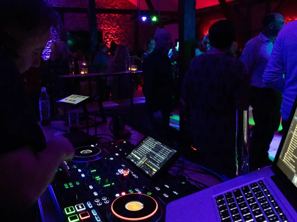 Ein DJ legt live auf, das Mischpult leuchtet bunt im Takt der Musik. Im Hintergrund füllen Gäste die Tanzfläche, stimmungsvolle Beleuchtung in Blau und Rot verleiht dem Raum Club-Atmosphäre. Ein Moment voller Rhythmus und Energie.