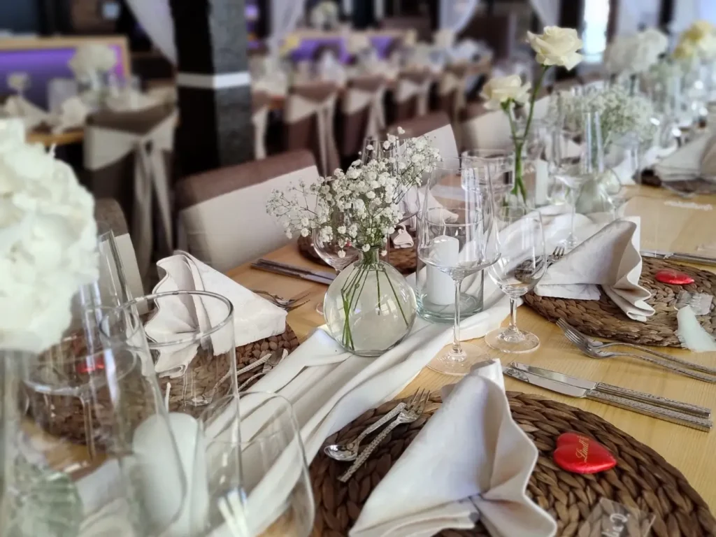 Lange Tafeln mit liebevoller Dekoration: Stoffservietten, stilvolle Gläser, schlichte Blumengestecke und rote Schokoherzen als persönliche Note. Ein stilvoll gedeckter Raum, bereit für eine Hochzeit oder ein festliches Dinner.