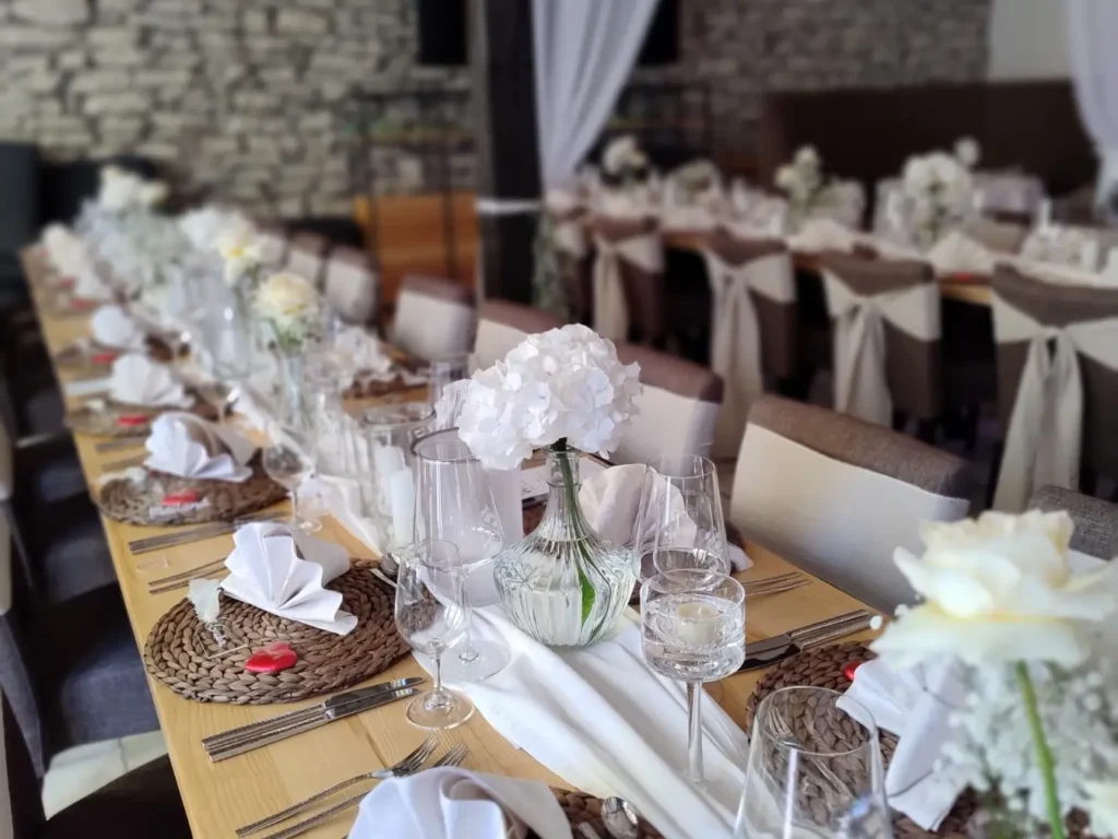 Ein Blick entlang einer langen, festlich gedeckten Tafel. Weiße Hortensien, Leinenservietten und elegante Glasvasen schmücken den Raum. Die Atmosphäre wirkt festlich, zugleich natürlich und stilvoll.
