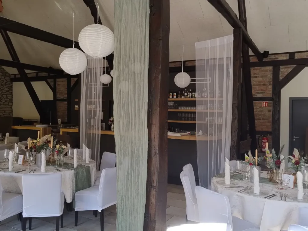 Der helle, rustikale Raum kombiniert Fachwerkcharme mit moderner Eleganz. Weiße Hussenstühle, runde Tische mit floraler Deko und große Papierlampions schaffen einen festlichen Rahmen – ideal für Trauungen, Geburtstage oder Firmenfeiern.
