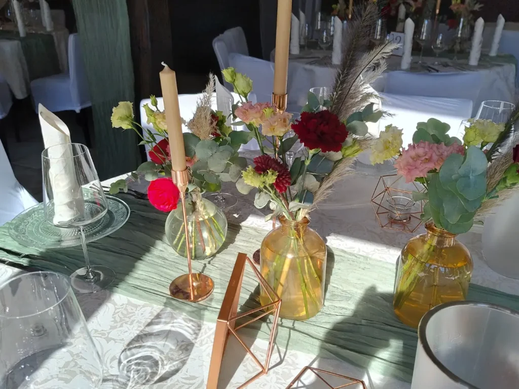 Zart arrangierte Blumen in goldfarbenen Vasen, kombiniert mit Kerzenhaltern und geometrischer Deko – ein liebevoll gestaltetes Detail für stilvolle Feste. Das Licht fällt sanft auf Tischdecke und Glas, und verleiht dem Setting Wärme und Eleganz.