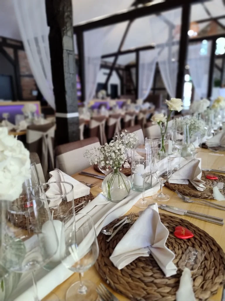 Festlich gedeckter Tisch mit geflochtenen Platzsets, weißen Servietten, Gläsern und kleinen Blumenvasen in rustikalem Saal mit Holzbalken.