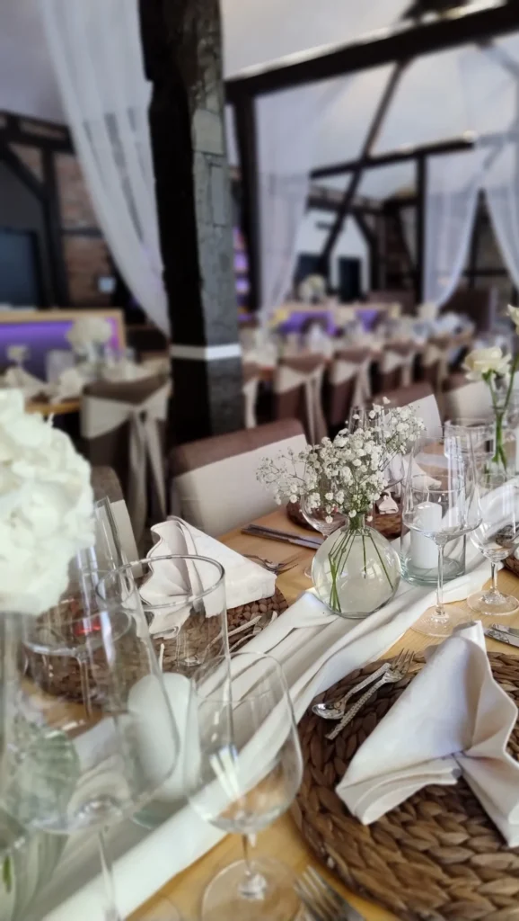 Festlich gedeckter Tisch mit geflochtenen Platzsets, weißen Servietten, Gläsern und kleinen Blumenvasen in rustikalem Saal mit Holzbalken.