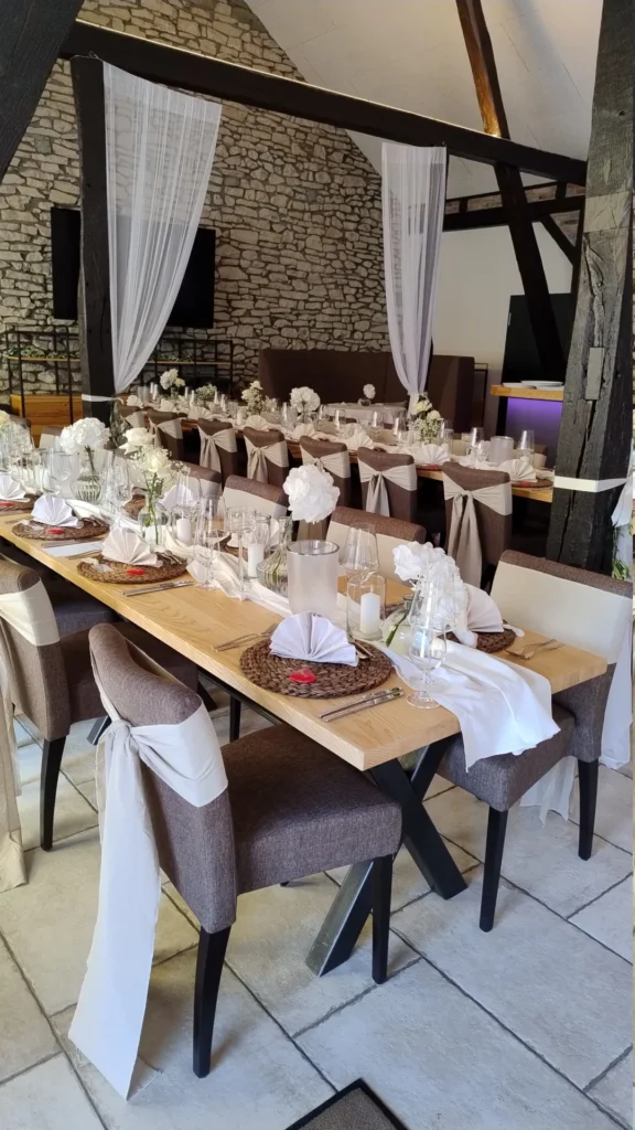 Langer festlich gedeckter Tisch mit weißen Servietten, Gläsern, Kerzen und Blumen in rustikalem Saal mit Steinwand.