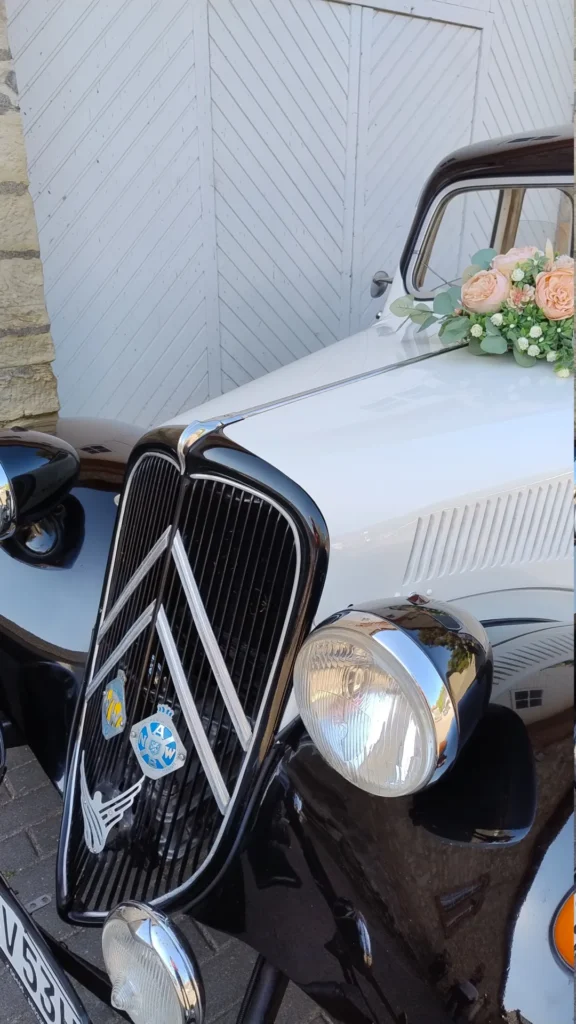 Nahaufnahme eines klassischen, schwarz-weißen Oldtimers mit floraler Dekoration auf der Motorhaube, geparkt vor einem hellen Holztor mit diagonaler Musterung.