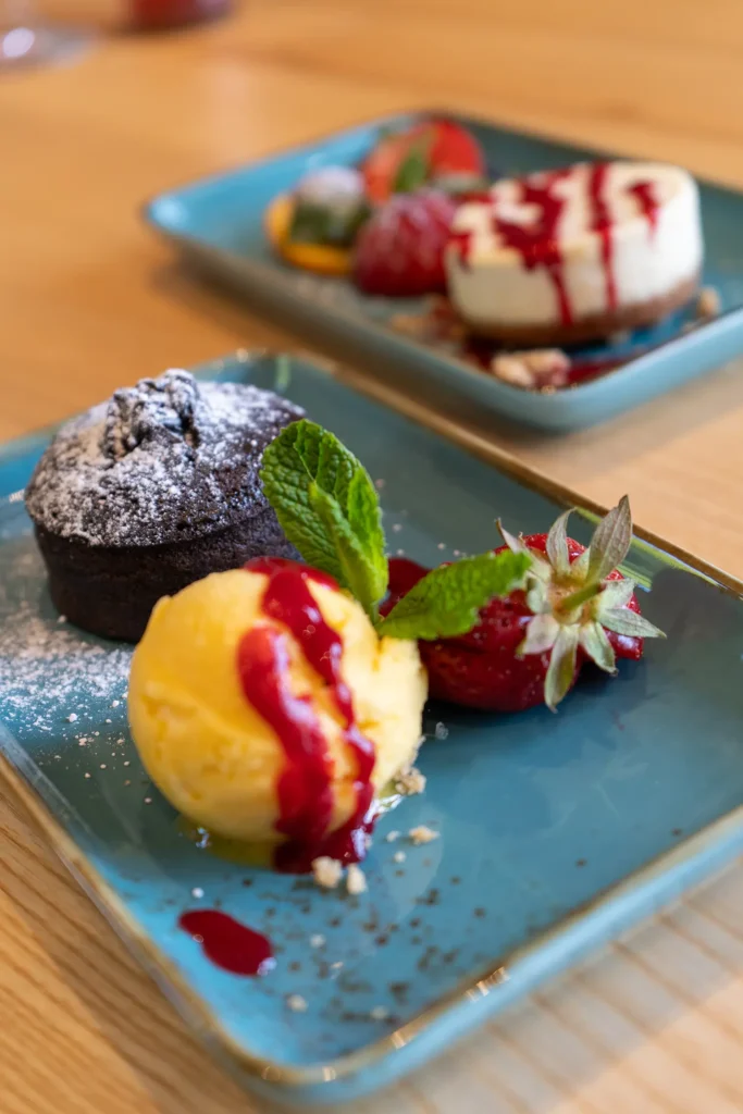 Ein Dessertteller mit einem warmen Schokoküchlein, bestäubt mit Puderzucker, serviert mit einer Kugel Vanilleeis, garniert mit Minze, frischen Erdbeeren und roter Fruchtsauce. Im Hintergrund ist unscharf ein zweiter Teller mit weiteren Desserts zu erkennen.