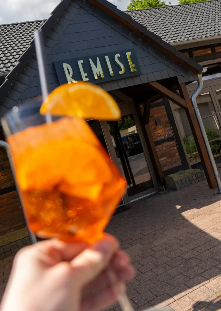 Im Vordergrund ist ein Glas mit Aperol Spritz zu sehen, dekoriert mit einer Orangenscheibe. Das Getränk wird unscharf gehalten, während im Hintergrund der Eingangsbereich eines Gebäudes mit dem Schild ‚Remise‘ gut lesbar ist. Der Eingang besteht aus einer dunklen Holzkonstruktion mit Backsteinfassade.
