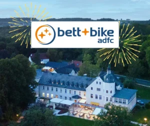 Luftaufnahme des abends beleuchteten Haus Delecke, umgeben von Bäumen und Natur. Im oberen Bildbereich ist das Logo „bett+bike adfc“ prominent eingeblendet, rechts und links davon stilisierte gelbe Feuerwerksstrahlen als grafisches Element zur Feier der Zertifizierung. Wenn du magst, kann ich auch eine Version in einfacher Sprache oder für Screenreader optimieren.