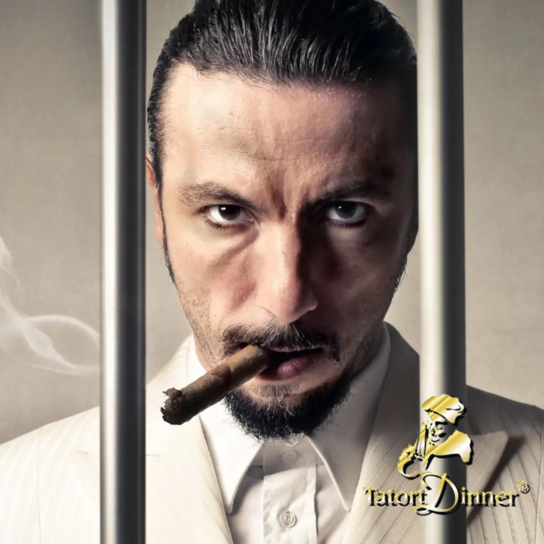 Ein Mann mit ernstem Gesichtsausdruck und streng zurückgekämmtem Haar hält einen Zigarillo im Mund und schaut direkt in die Kamera. Er trägt einen eleganten Anzug und steht hinter Gefängnisgittern. Der Hintergrund ist neutral gehalten und erzeugt eine dramatische Atmosphäre. In der unteren rechten Ecke des Bildes befindet sich das Logo von "Tatort