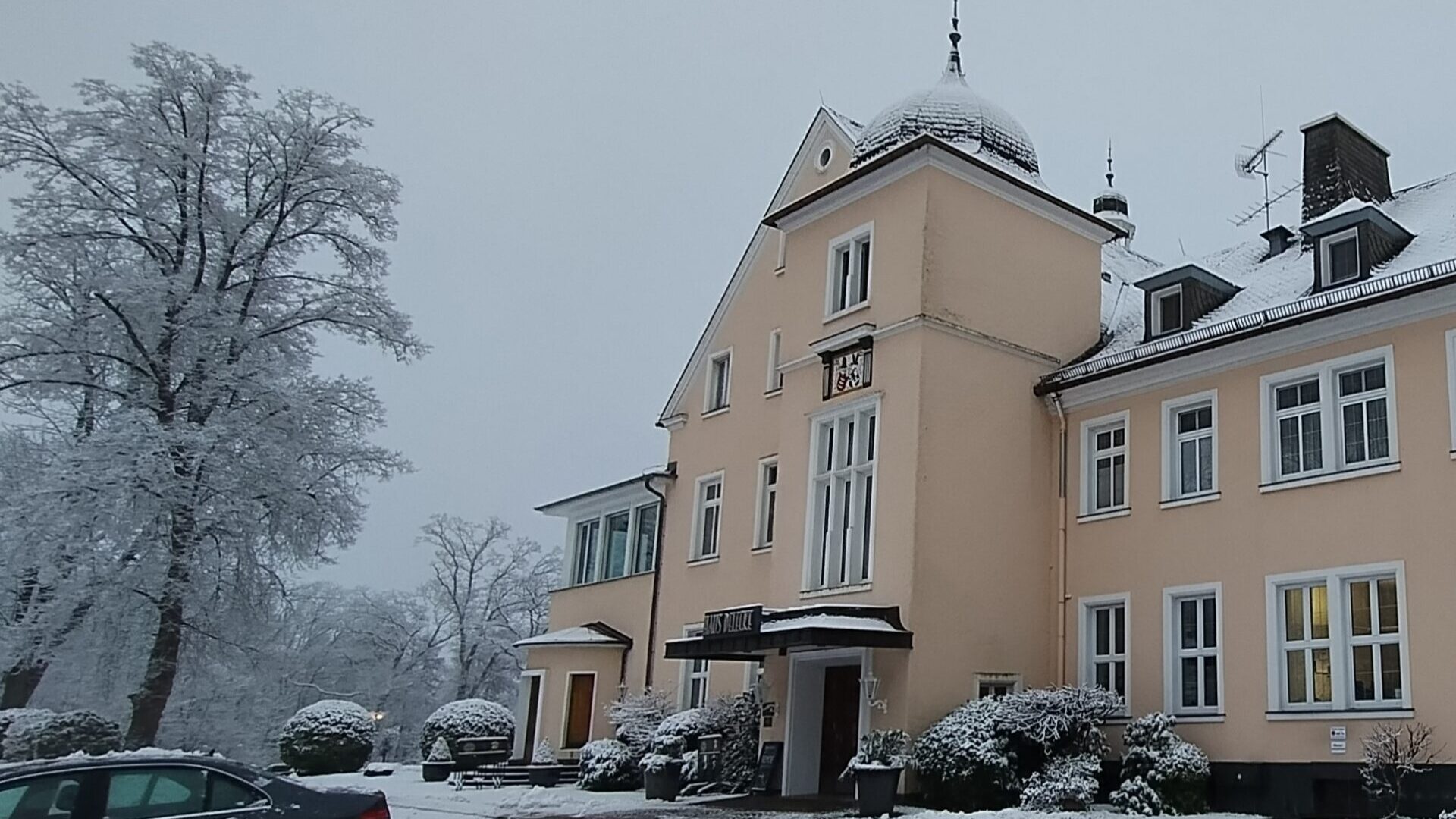 Alt-Text: Winterliche Ansicht eines zweigeschossigen, hellgelben Gebäudes mit einem kuppelförmigen Dach und großen Fenstern. Der Eingangsbereich ist mit Schnee bedeckt, und im Vordergrund sind schneebedeckte Bäume und ein geparktes Auto zu sehen. Der Himmel ist grau und bewölkt.